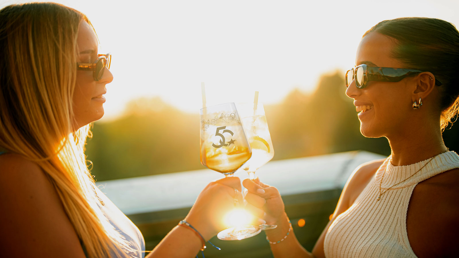 Zwei junge Damen trinken im Sonnenuntergang aus den Jubiläums-Weingläsern. 